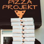 A person carrying several pizza boxes stacked high with the Pizza Projekt logo and front door behind.